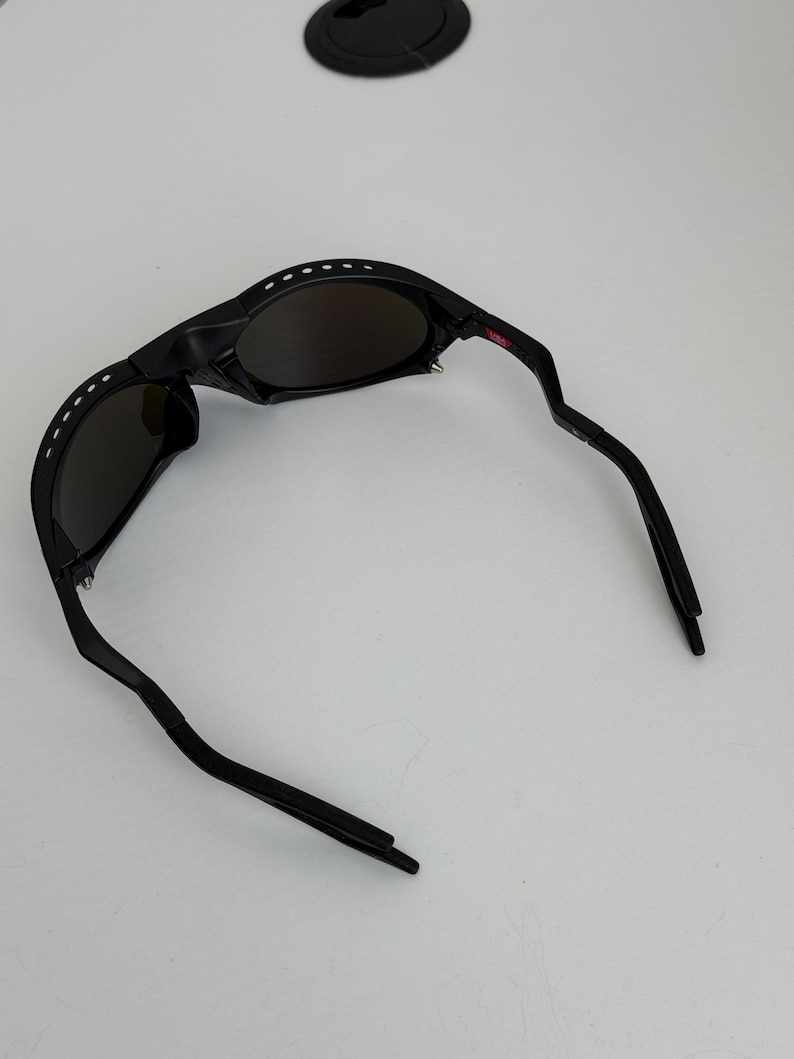 May include: Black sunglasses with a curved frame and dark lenses. The sunglasses have a series of white dots along the top of the frame and a red accent on the right arm. The arms are curved and angled.