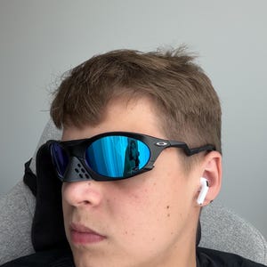 May include: Black sports sunglasses with blue mirrored lenses. The sunglasses have a distinctive design with a nose bridge and a sleek, modern frame. The lenses reflect a blue hue, and the frame is black.