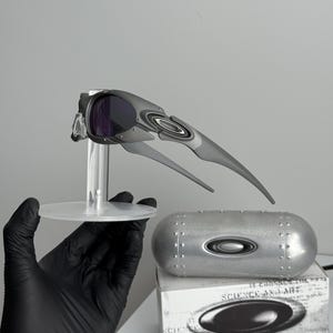 May include: Gray and black Oakley sunglasses with purple lenses are displayed on a clear stand. A silver case and a white box with the text "SCIENCE AND ART" are also visible. The sunglasses have a futuristic design.
