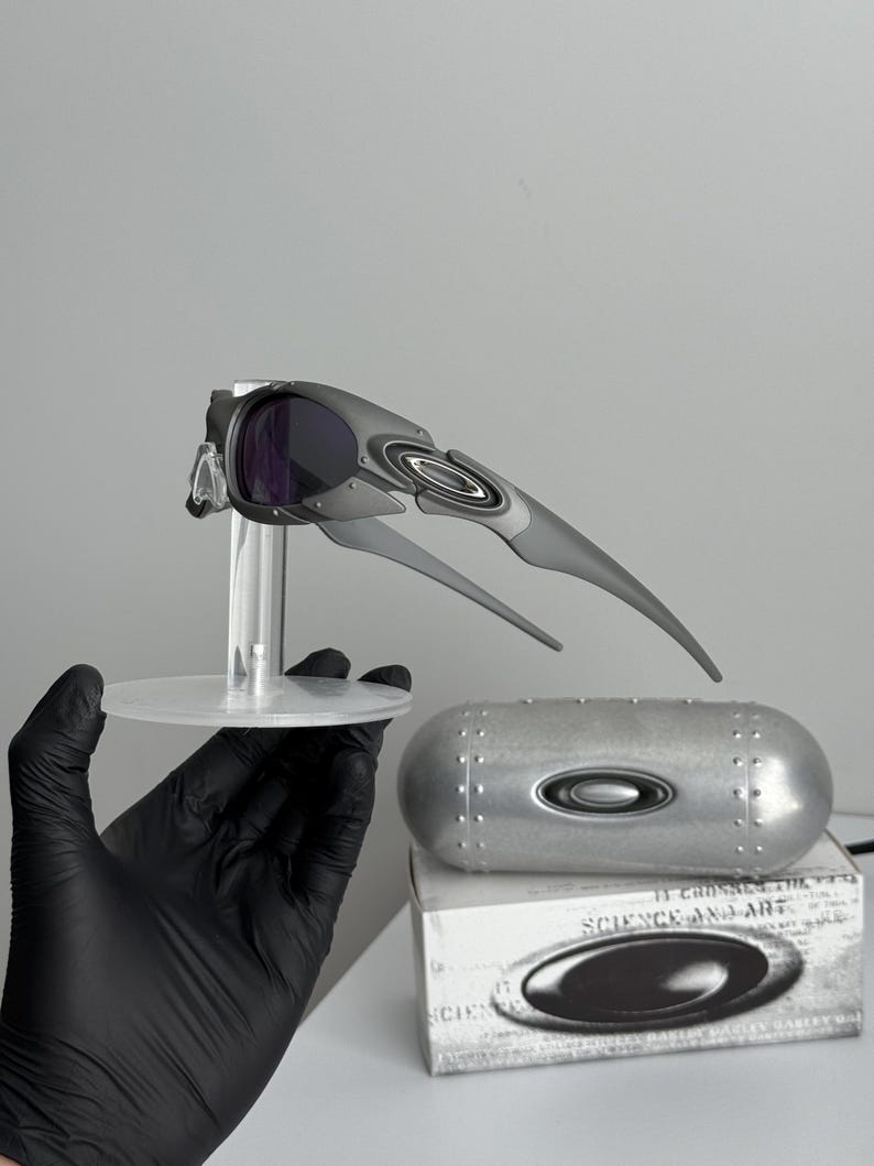 May include: Gray and black Oakley sunglasses with purple lenses are displayed on a clear stand. A silver case and a white box with the text "SCIENCE AND ART" are also visible. The sunglasses have a futuristic design.