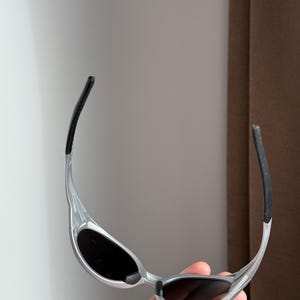 May include: Silver sunglasses with black lenses and black temple tips. The sunglasses have a sleek, modern design with a curved frame and a minimalist aesthetic. The lenses are oval-shaped.