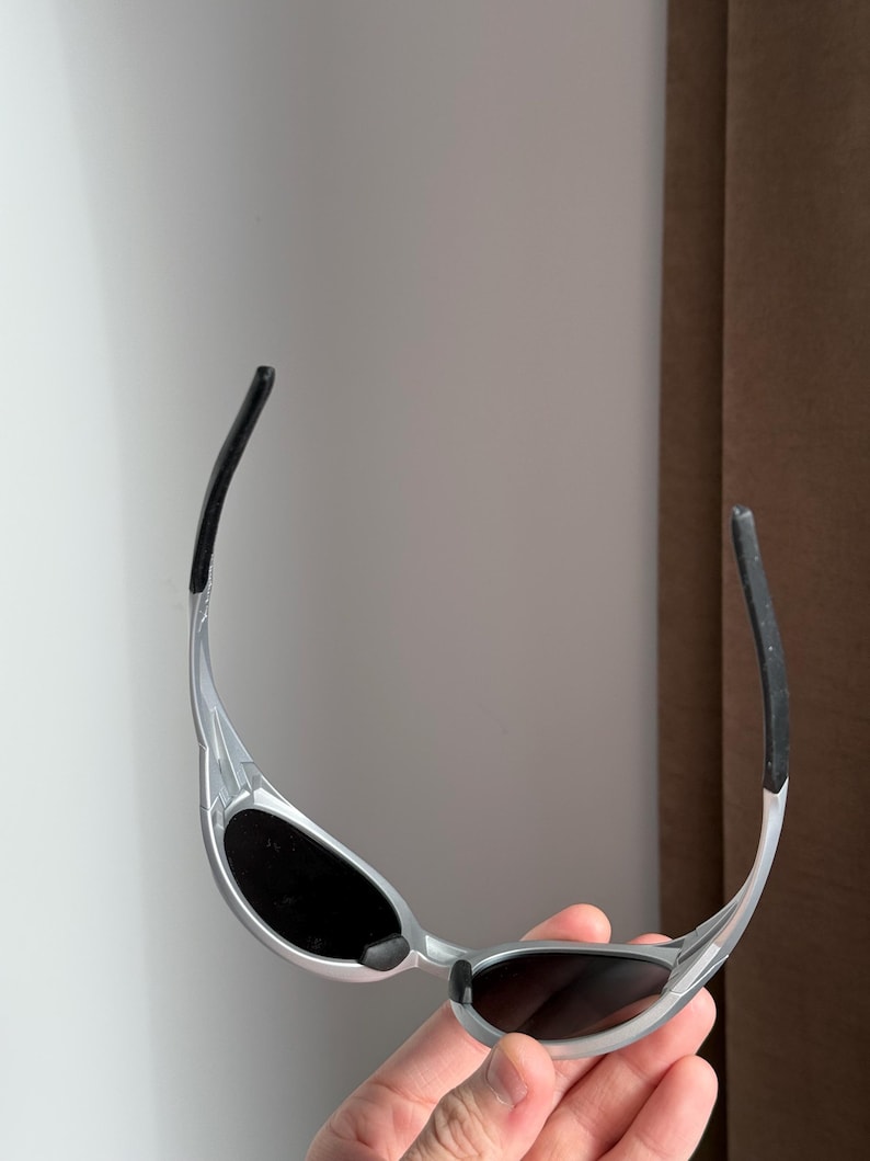 May include: Silver sunglasses with black lenses and black temple tips. The sunglasses have a sleek, modern design with a curved frame and a minimalist aesthetic. The lenses are oval-shaped.