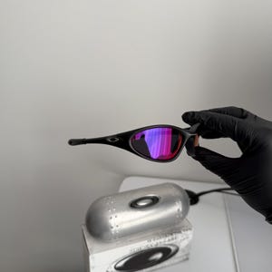 May include: Black sunglasses with a curved frame and mirrored lenses that reflect shades of pink and purple. The sunglasses are held by a gloved hand, with a silver case and box in the background.
