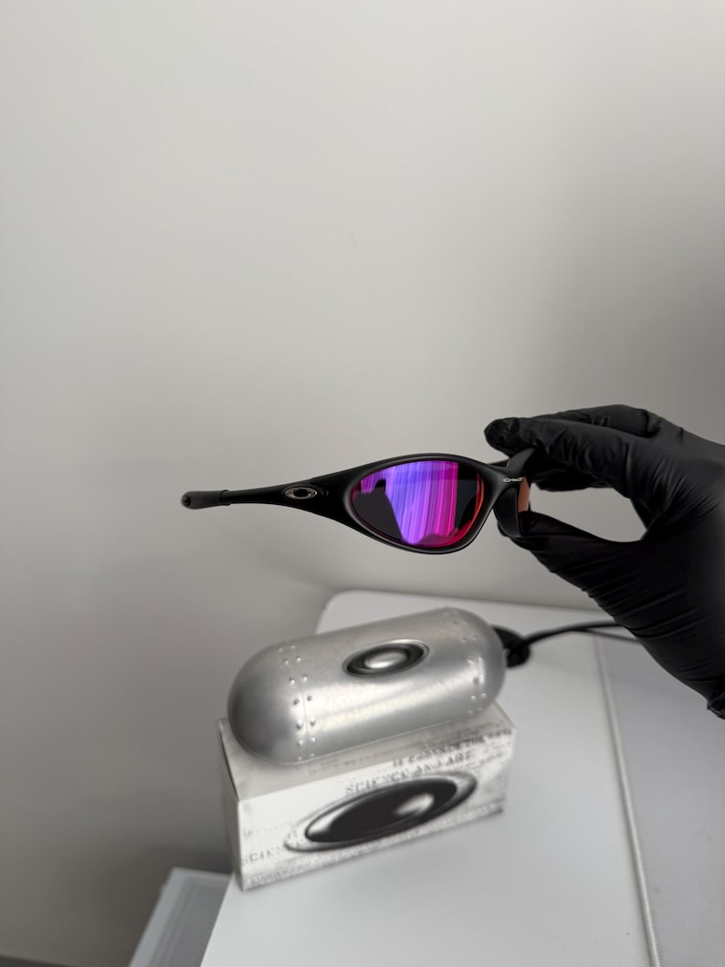 May include: Black sunglasses with a curved frame and mirrored lenses that reflect shades of pink and purple. The sunglasses are held by a gloved hand, with a silver case and box in the background.