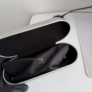 May include: Open silver-colored Oakley glasses case with a black interior and a black microfiber cloth. The Oakley logo is visible on the cloth. The case is open, revealing the contents.