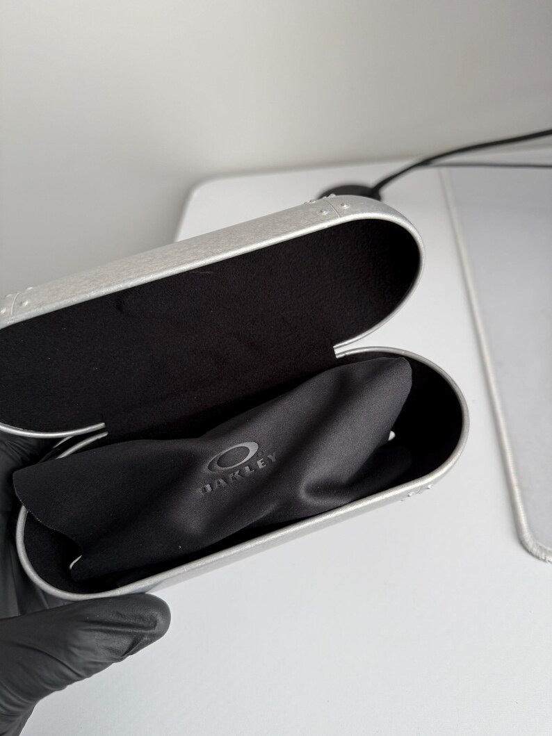 May include: Open silver-colored Oakley glasses case with a black interior and a black microfiber cloth. The Oakley logo is visible on the cloth. The case is open, revealing the contents.