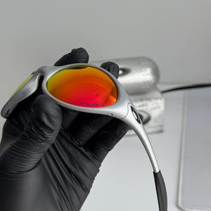 Pu&ograve; includere: Occhiali da sole argentati con lenti rosso-arancio vivaci. Gli occhiali hanno un design elegante e moderno con aste curve e terminali neri. Le lenti sono riflettenti e la montatura ha una finitura metallica.