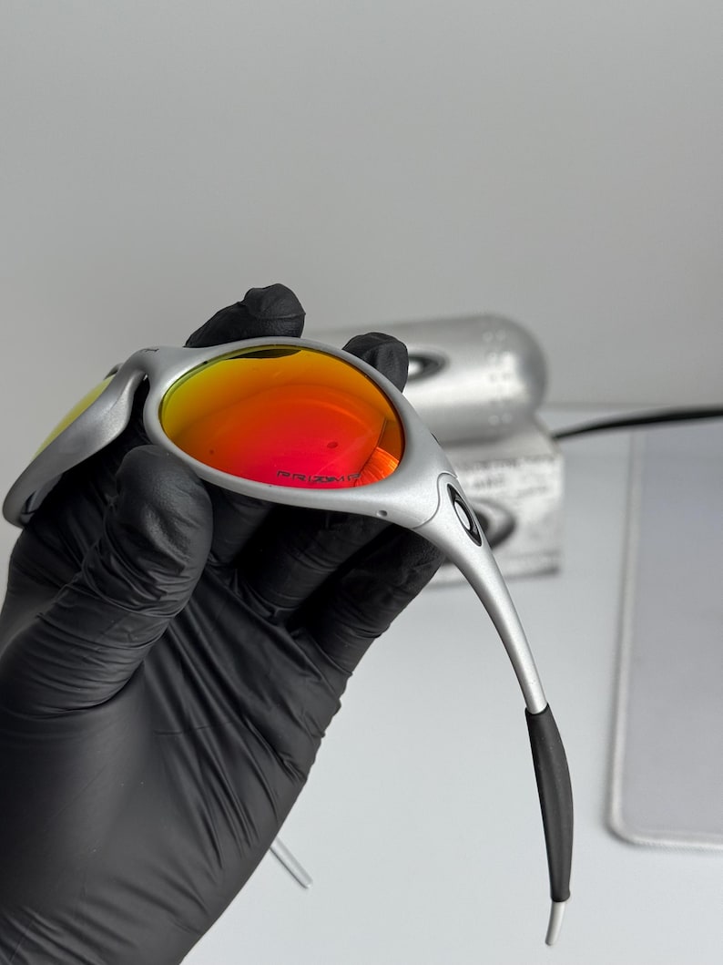 Pu&ograve; includere: Occhiali da sole argentati con lenti rosso-arancio vivaci. Gli occhiali hanno un design elegante e moderno con aste curve e terminali neri. Le lenti sono riflettenti e la montatura ha una finitura metallica.