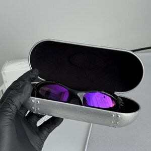 May include: Black sunglasses with purple mirrored lenses rest inside a silver case with a black interior. The sunglasses have a sleek, modern design. The case is open, revealing the glasses.