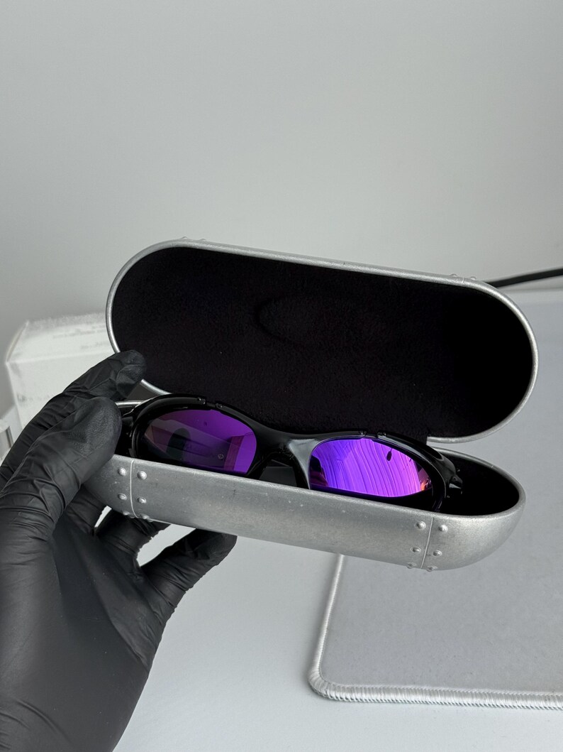 May include: Black sunglasses with purple mirrored lenses rest inside a silver case with a black interior. The sunglasses have a sleek, modern design. The case is open, revealing the glasses.