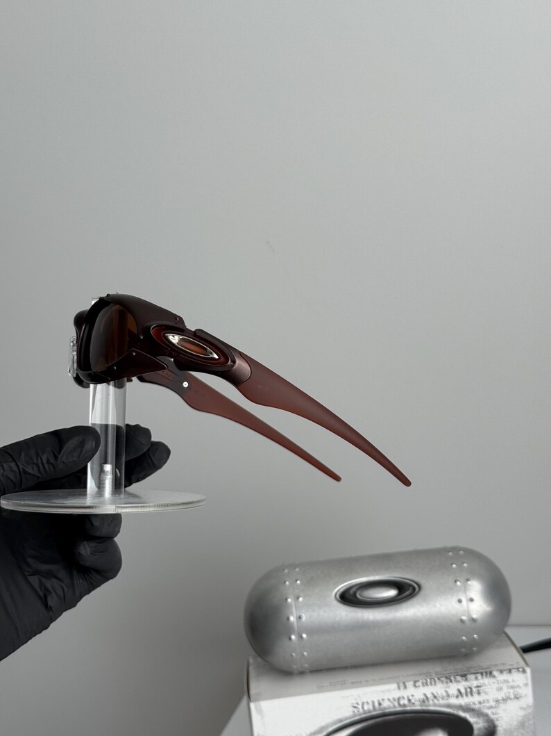 May include: Brown and red sunglasses with a unique, angular design. The sunglasses are held on a clear stand, with a silver case and a box visible in the background. The case has the Oakley logo.