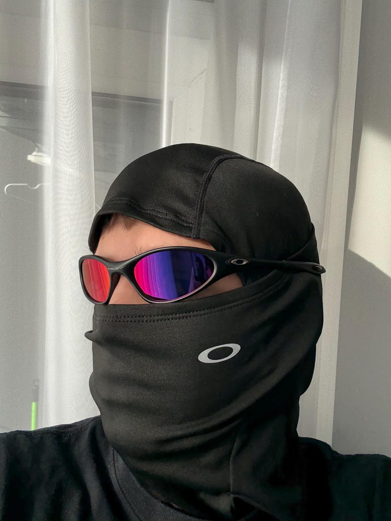 May include: Black balaclava with a white Oakley logo and black sunglasses with red and purple lenses. The sunglasses have a black frame. The balaclava covers the head and neck.
