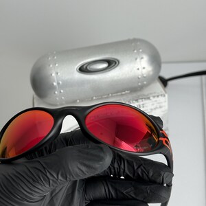 May include: Black sunglasses with red lenses and a flame design on the temples. The sunglasses are held in a black gloved hand. A silver case with the Oakley logo is in the background.