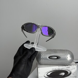 May include: Gray sunglasses with purple lenses are displayed on a clear stand. A silver case and a white box with the text "SCIENCE AND ART" are also visible. The sunglasses have a sporty design.