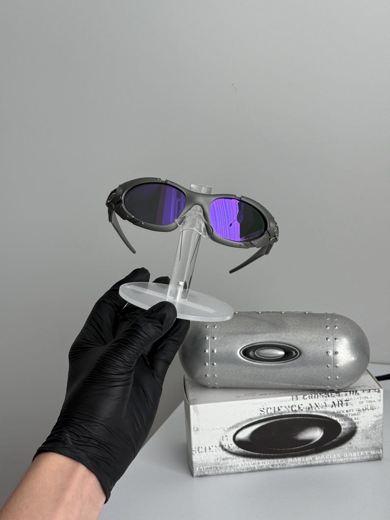 May include: Gray sunglasses with purple lenses are displayed on a clear stand. A silver case and a white box with the text "SCIENCE AND ART" are also visible. The sunglasses have a sporty design.
