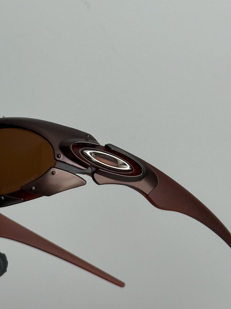 May include: Close-up of brown sunglasses with a metallic frame. The sunglasses have a curved design with a silver accent near the hinge and brown tinted lenses. The arms of the sunglasses are a darker brown.