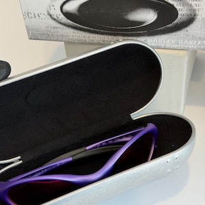 May include: Purple sunglasses with a matching case. The sunglasses have a sleek, modern design with a purple frame and dark lenses. The case is silver with a black interior. The box behind the case has the words "SCIENCE AND ART".