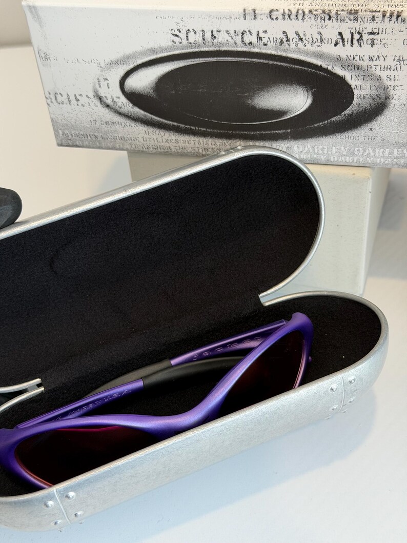 May include: Purple sunglasses with a matching case. The sunglasses have a sleek, modern design with a purple frame and dark lenses. The case is silver with a black interior. The box behind the case has the words "SCIENCE AND ART".