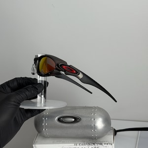 May include: Black and silver sunglasses with red and yellow lenses. The sunglasses are displayed on a clear stand, with a silver case and a box with the text "SCIENCE AND ART" visible in the background.