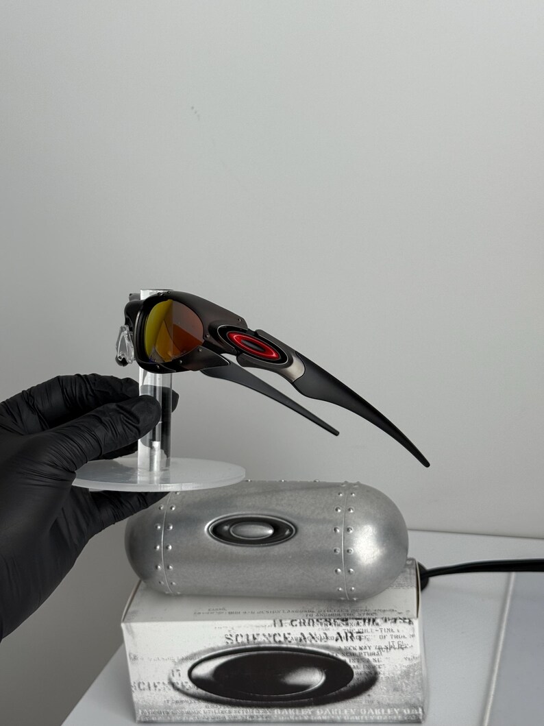 May include: Black and silver sunglasses with red and yellow lenses. The sunglasses are displayed on a clear stand, with a silver case and a box with the text "SCIENCE AND ART" visible in the background.