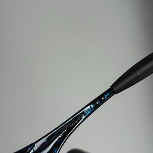 May include: Close-up of black sunglasses with a blue speckled design on the frame. The text "MADE IN" is visible on the arm of the glasses. The sunglasses have a sleek, modern design.