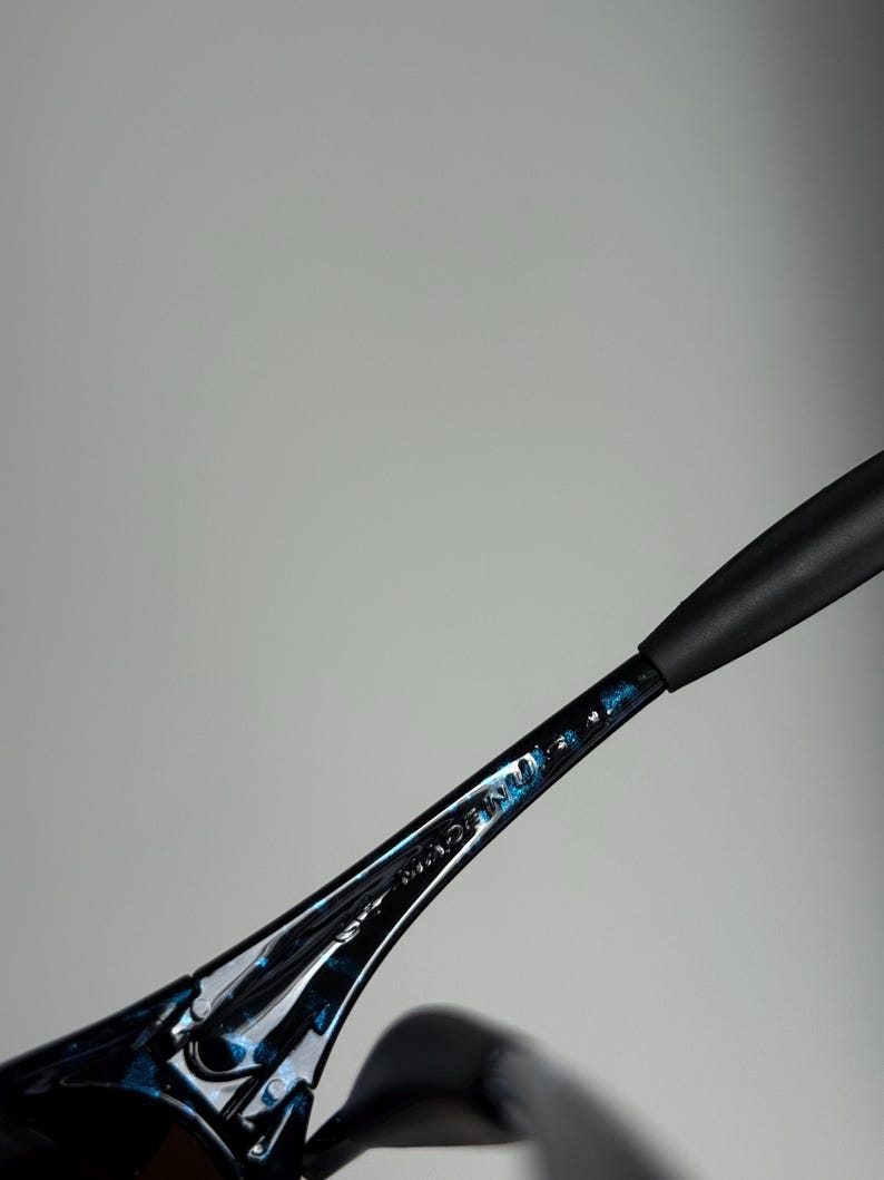 May include: Close-up of black sunglasses with a blue speckled design on the frame. The text "MADE IN" is visible on the arm of the glasses. The sunglasses have a sleek, modern design.