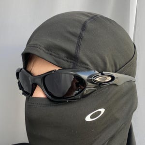 May include: Black sunglasses with a curved frame and dark lenses. The sunglasses are worn over a black balaclava. The Oakley logo is visible on the temple and the balaclava.