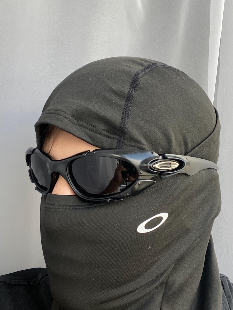 May include: Black sunglasses with a curved frame and dark lenses. The sunglasses are worn over a black balaclava. The Oakley logo is visible on the temple and the balaclava.