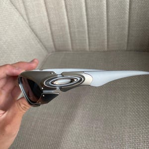 May include: A pair of sporty sunglasses with a white frame and dark lenses. The sunglasses have a silver metal accent on the temples and a sleek, modern design. The lenses appear to be tinted, offering protection from the sun.