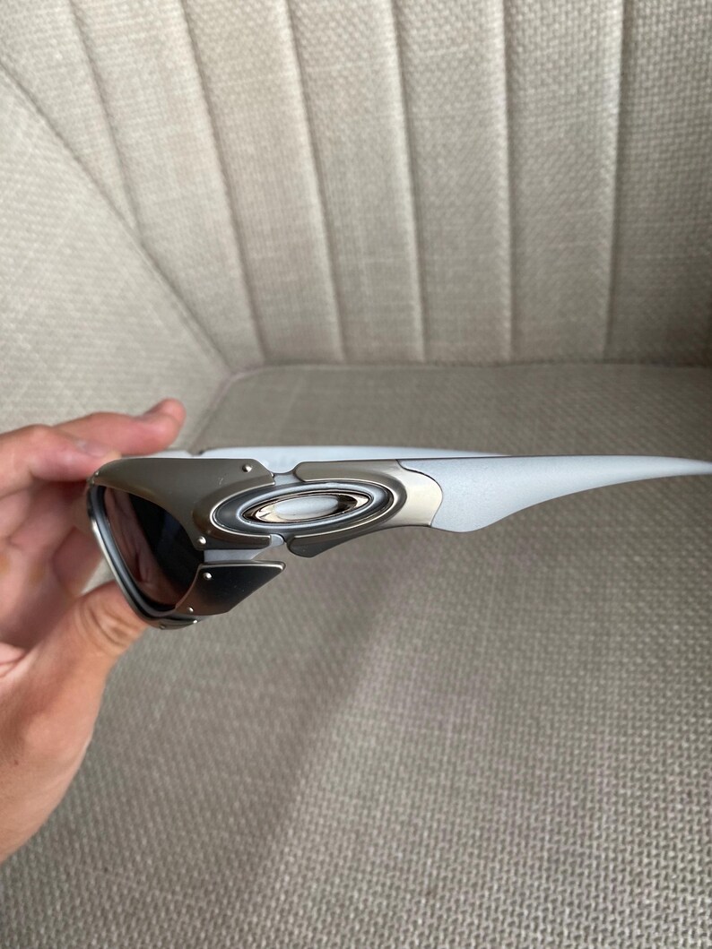 May include: A pair of sporty sunglasses with a white frame and dark lenses. The sunglasses have a silver metal accent on the temples and a sleek, modern design. The lenses appear to be tinted, offering protection from the sun.