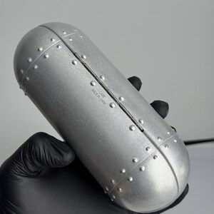 May include: A silver-colored, oval-shaped case with a riveted design. The case is held by a gloved hand. The text "CASE MADE IN CHINA" is printed on the case.