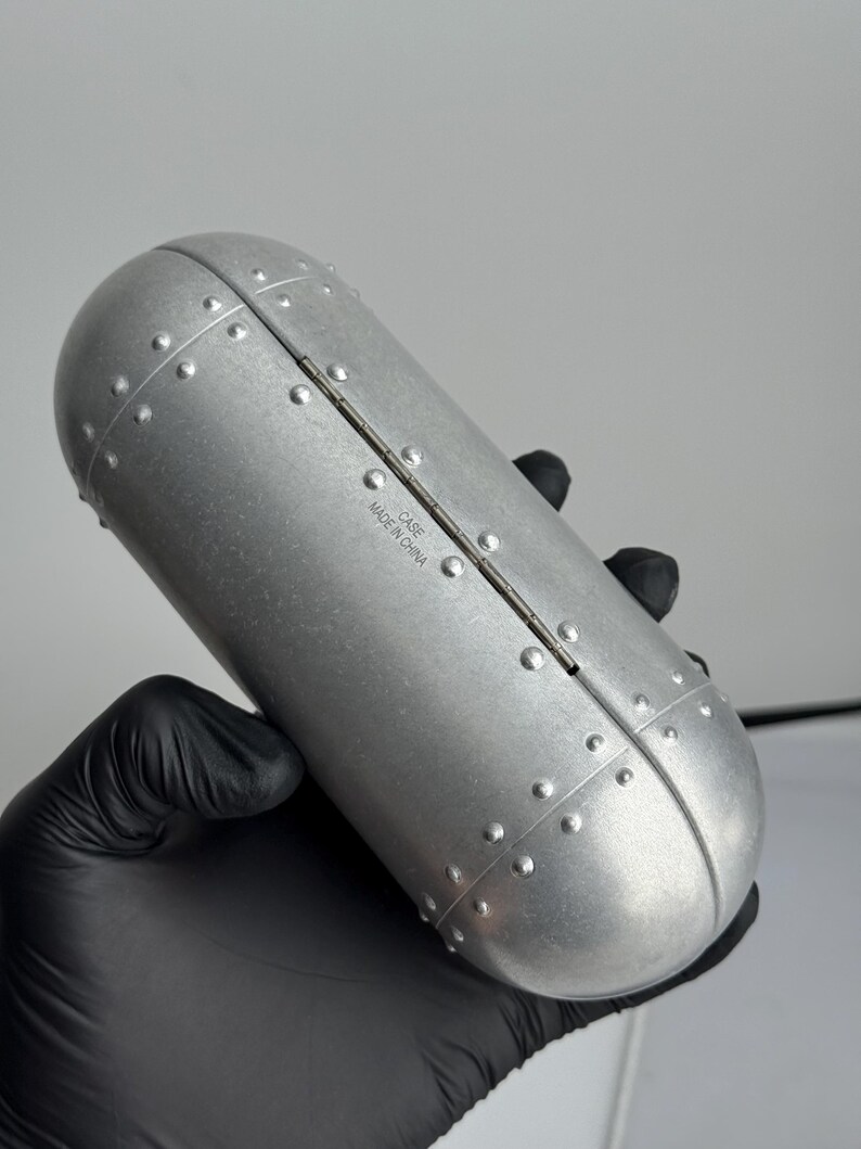 May include: A silver-colored, oval-shaped case with a riveted design. The case is held by a gloved hand. The text "CASE MADE IN CHINA" is printed on the case.
