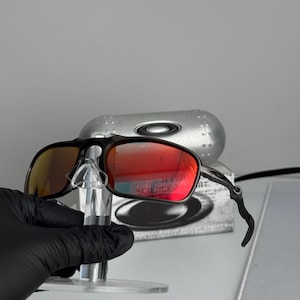 May include: Black framed sunglasses with red-tinted lenses. The sunglasses are displayed on a clear stand, with a silver case in the background. The lenses reflect a bright orange and red hue.