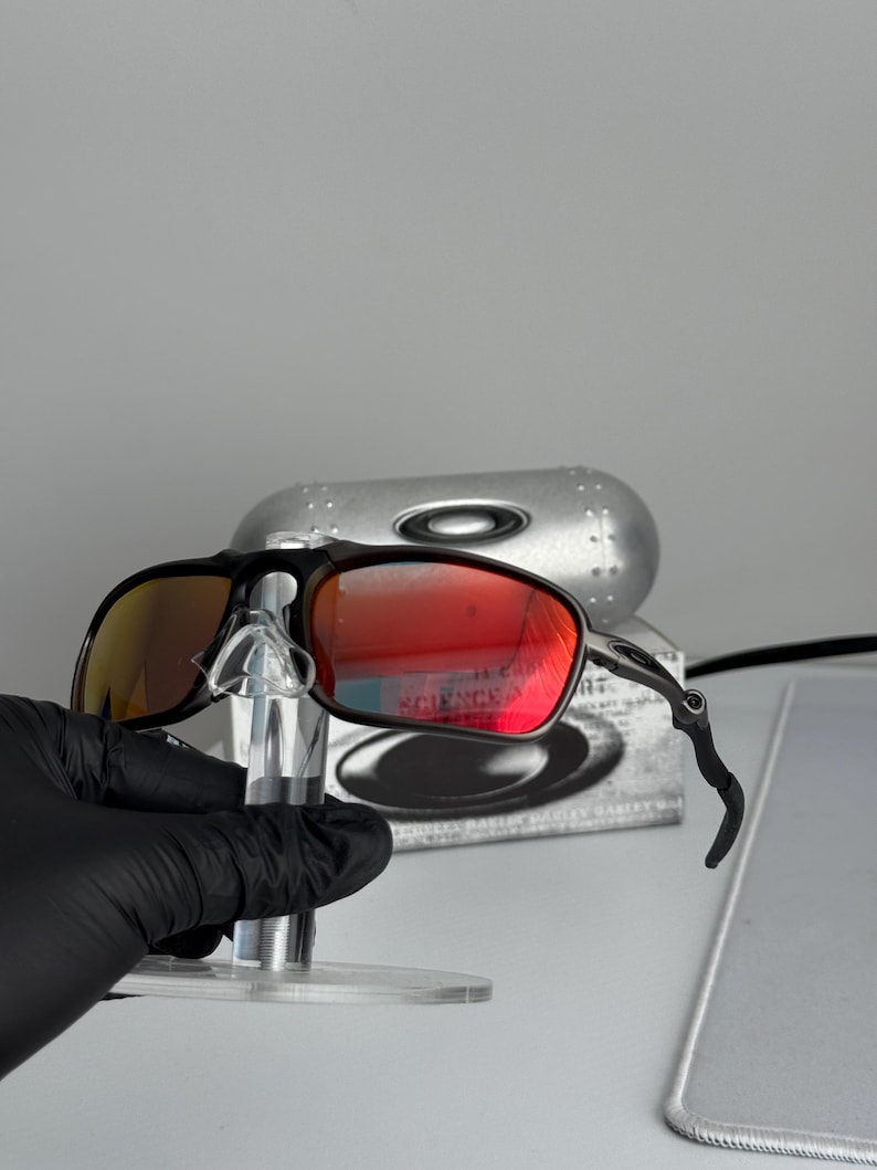 May include: Black framed sunglasses with red-tinted lenses. The sunglasses are displayed on a clear stand, with a silver case in the background. The lenses reflect a bright orange and red hue.