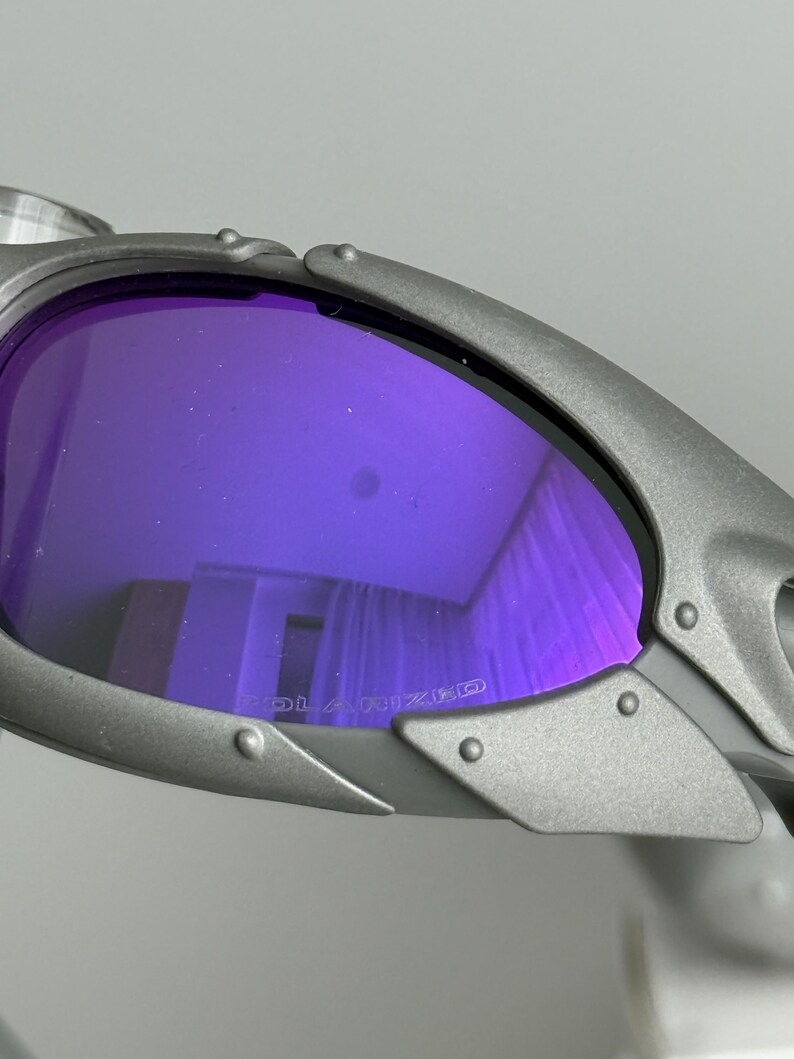 May include: Close-up of gray sunglasses with purple polarized lenses. The lenses reflect the surrounding environment. The frame has a riveted design. The word "POLARIZED" is printed on the lens.