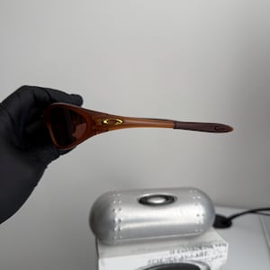 May include: Brown sunglasses with a matching case and box. The sunglasses have a brown frame and lenses. The case is silver and oval-shaped. The box has the text "SCIENCE AND ART" and the Oakley logo.