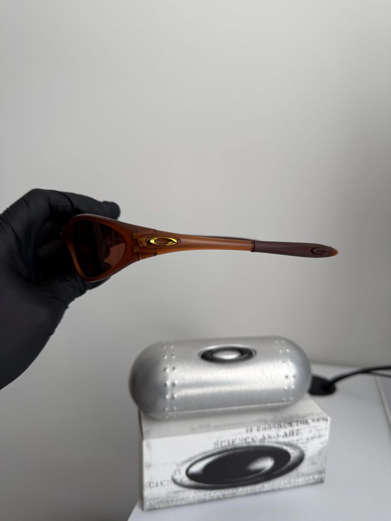 May include: Brown sunglasses with a matching case and box. The sunglasses have a brown frame and lenses. The case is silver and oval-shaped. The box has the text "SCIENCE AND ART" and the Oakley logo.