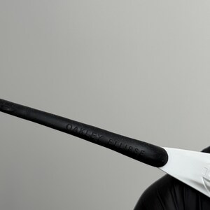 Puede incluir: Primer plano de gafas de sol Oakley Eclipse. La patilla negra de las gafas tiene el texto "OAKLEY ECLIPSE" impreso. La montura es negra y blanca.