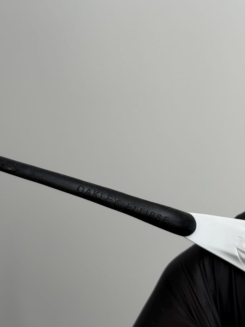 Puede incluir: Primer plano de gafas de sol Oakley Eclipse. La patilla negra de las gafas tiene el texto "OAKLEY ECLIPSE" impreso. La montura es negra y blanca.