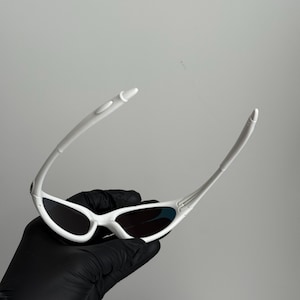May include: White sunglasses with a streamlined design. The sunglasses feature a white frame and dark lenses. The arms are slender and curve. The sunglasses are held by a gloved hand. A contemporary eyewear accessory.