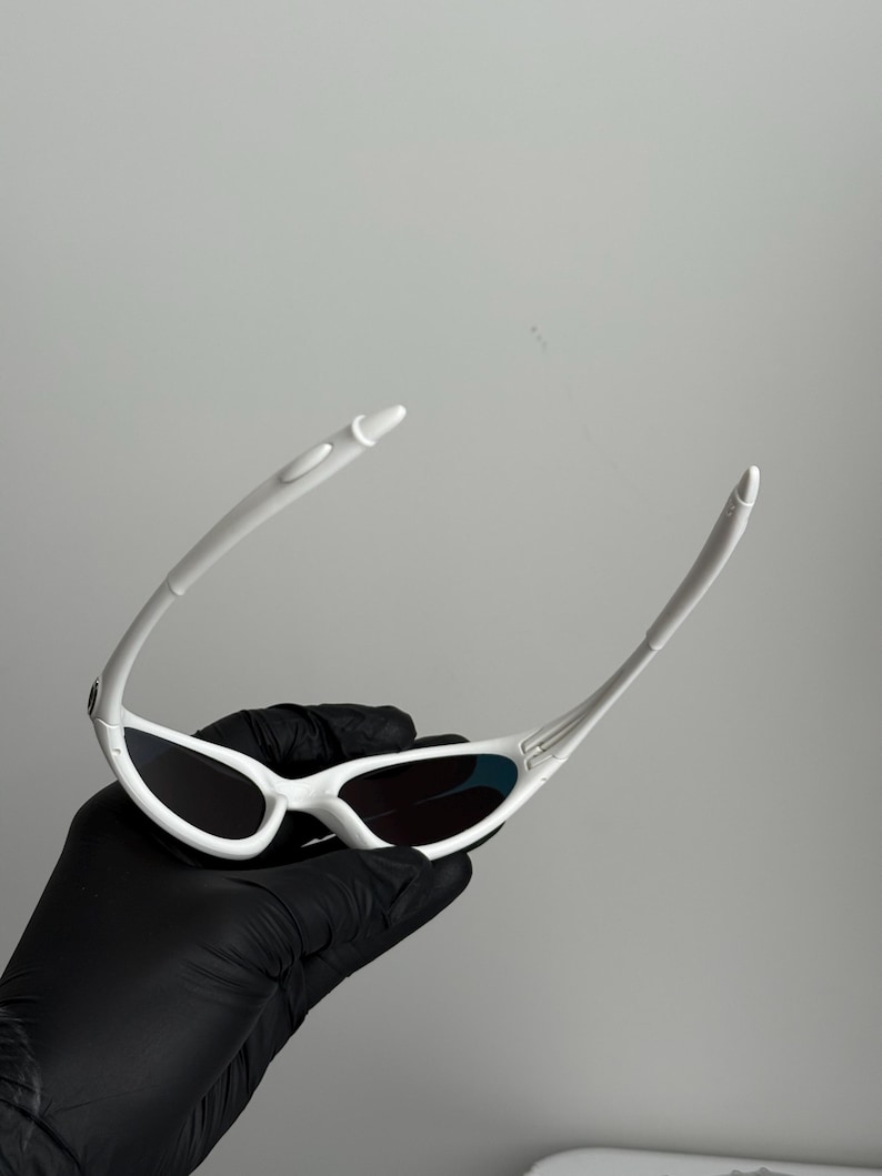 May include: White sunglasses with a streamlined design. The sunglasses feature a white frame and dark lenses. The arms are slender and curve. The sunglasses are held by a gloved hand. A contemporary eyewear accessory.