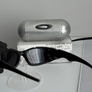 May include: Black Oakley sunglasses with a silver case. The sunglasses have a rectangular frame and dark lenses. The case is silver with a black Oakley logo and a riveted design.