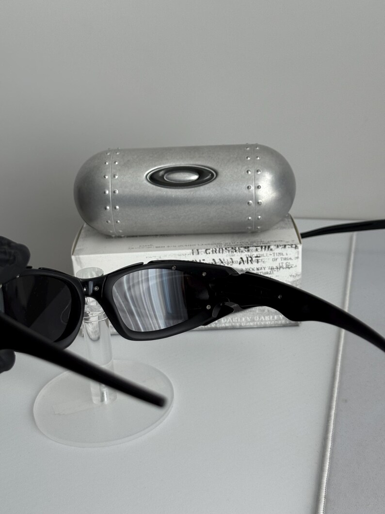 May include: Black Oakley sunglasses with a silver case. The sunglasses have a rectangular frame and dark lenses. The case is silver with a black Oakley logo and a riveted design.
