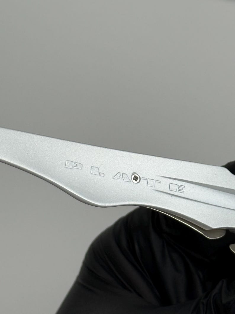 May include: A close-up of a silver metal tool with etched markings. The tool has a curved blade and a series of small, geometric symbols etched into its surface. The tool is held by a person wearing a black glove.