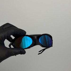 May include: Black sports sunglasses with blue mirrored lenses. The sunglasses have a curved frame and a sleek design. The lenses reflect a vibrant blue color. The sunglasses are held by a gloved hand.