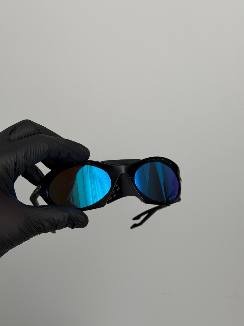 May include: Black sports sunglasses with blue mirrored lenses. The sunglasses have a curved frame and a sleek design. The lenses reflect a vibrant blue color. The sunglasses are held by a gloved hand.