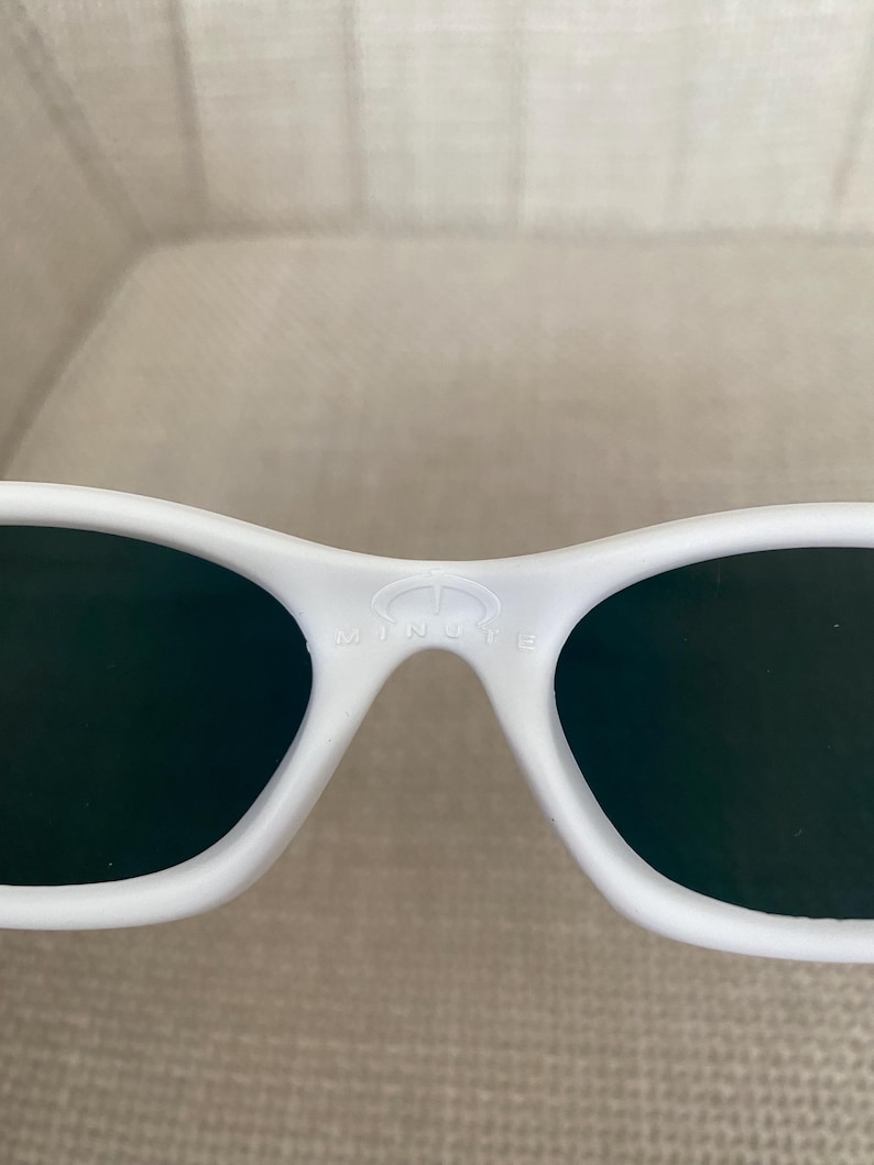 Puede incluir: Gafas 3D blancas con lentes oscuras. Las gafas tienen un dise&ntilde;o curvo y la palabra "MINUTE" est&aacute; impresa en el puente. Las gafas est&aacute;n hechas de un material liso y brillante.