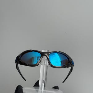 May include: Black framed sunglasses with blue mirrored lenses. The sunglasses are displayed on a clear plastic stand. The lenses reflect the surrounding environment, creating a vibrant blue hue.