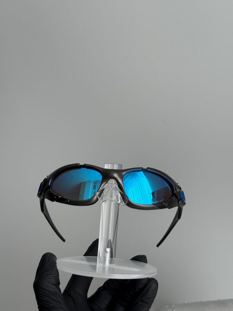 May include: Black framed sunglasses with blue mirrored lenses. The sunglasses are displayed on a clear plastic stand. The lenses reflect the surrounding environment, creating a vibrant blue hue.