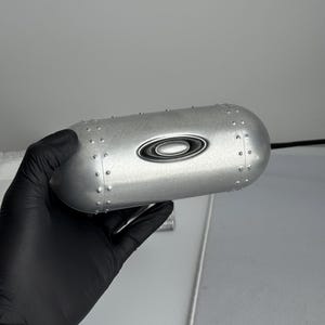 May include: A silver-colored Oakley case with a rounded, cylindrical shape. The case features the Oakley logo and small rivet details. The case is held by a gloved hand.